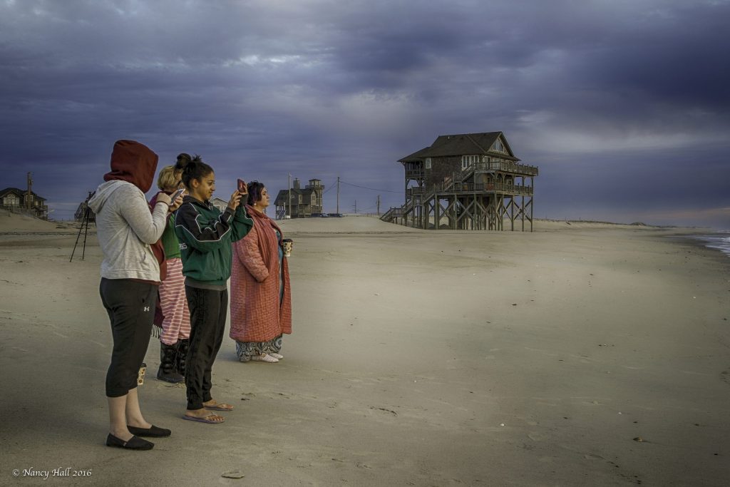 Four women stand on the beach. Two are using cell phones and the other two are looking out over the ocean