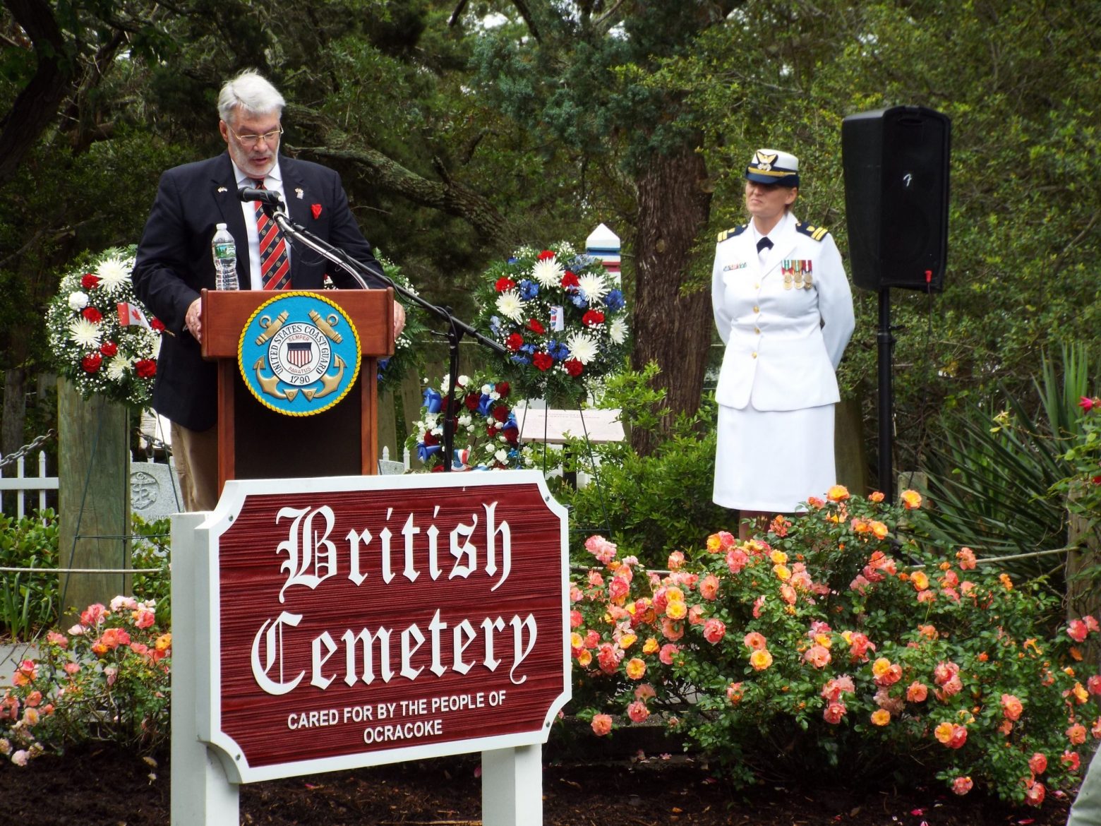 a man in a suit stands at a podium with a woman in a Navy uniform behind and to his left looking on. sign in front reads british cemetery
