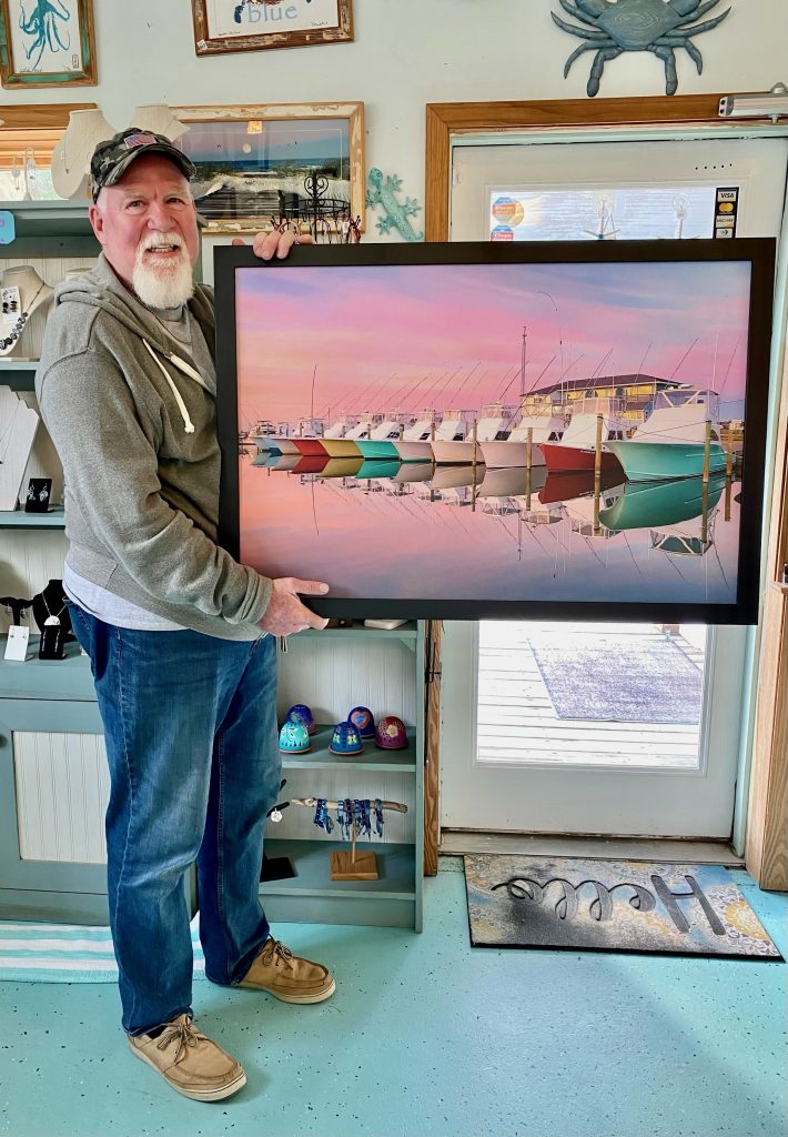 Man with white goatee wearing ballcap faces camera and holds a photograph of a line of brightly colored boats with a pink sky reflected on the calm water